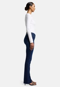 Jeans a zampa d'elefante in denim blu scuro con vita media, indossati con una camicia aderente bianca a maniche lunghe e sandali neri con cinturini.