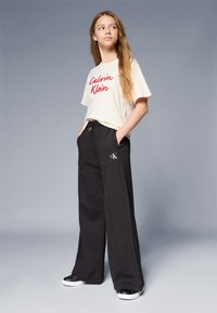 Bežna majica središnjega dela z rdečim besedilom "Calvin Klein", v kombinaciji z črnimi širokimi trenirkami, ki imajo majhen CK logo, ter črnimi športnimi čevlji.