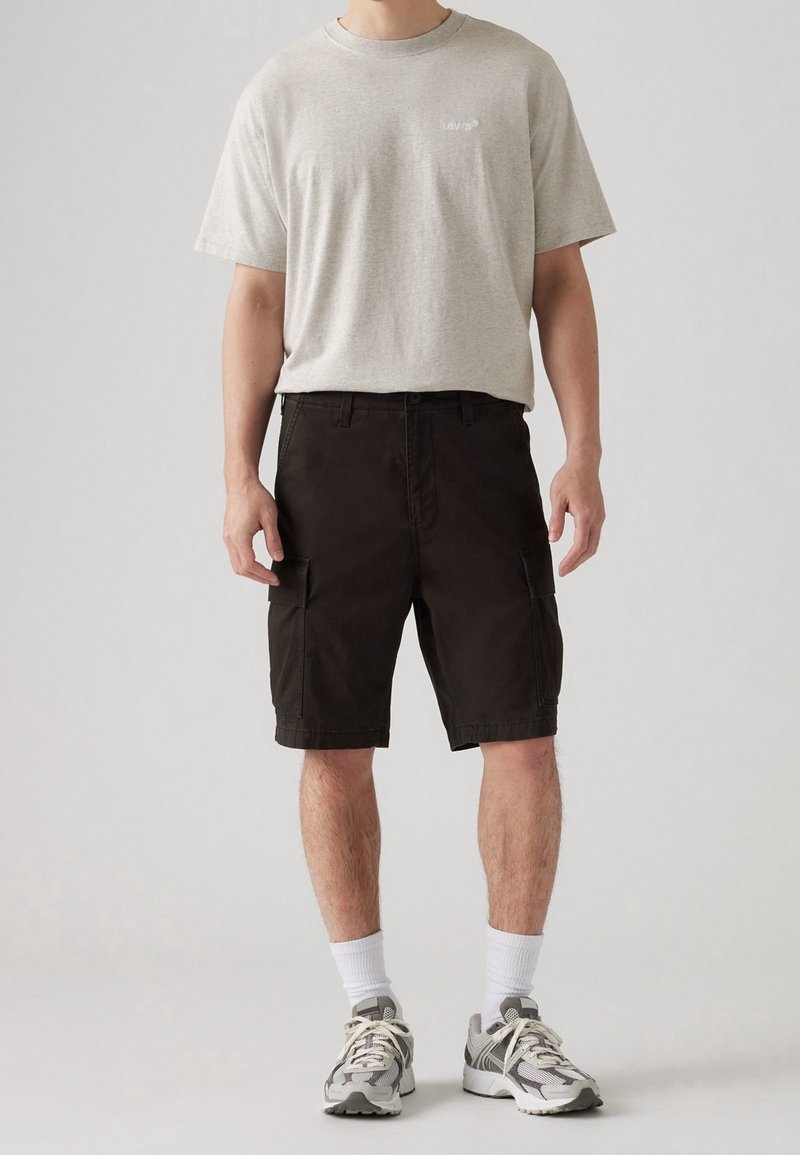 Homme portant un t-shirt gris clair à manches courtes, un short cargo noir aux genoux, des chaussettes blanches et des chaussures de course grises, debout devant un fond clair.
