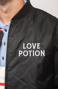 Black bomber jacket with a ribbed collar; features "LOVE POTION" embroidered in white on the front. Underneath is a light blue collared shirt.