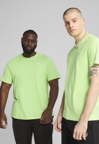 Doi bărbați îmbrăcați în tricouri cu mânecă scurtă, de culoare verde lime, din bumbac, cu mici logo-uri albe pe piept, asortați cu pantaloni negri.