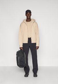 Felpa con zip beige a coste sopra una maglietta beige, abbinata a pantaloni neri e uno zaino nero, indossata con sneakers nere e un cappellino.
