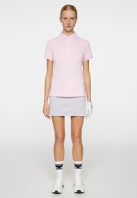 Lyserød polo-shirt med korte ærmer, tre knapper, parret med en grå nederdel. Modellen har hvide handsker og sortstribede strømper på.