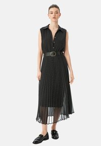 Schwarzes ärmelloses Kleid mit Kragen, mit durchgehender Knopfleiste, plissiertem Design und einem schwarzen Gürtel mit goldener Schnalle, getragen mit schwarzen Mokassins.