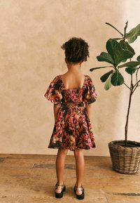 Buntes Blumenkleid mit Puffärmeln, gerafftem Oberteil und gestuftem Rock. Ein Paar schwarze Schuhe mit goldenen Akzenten und eine Pflanze im Hintergrund.