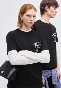 Δύο νέοι άνθρωποι στέκονται δίπλα δίπλα, φορώντας μαύρα μπλουζάκια με σχέδια. Η γυναίκα έχει σταυρωμένα τα χέρια κρατώντας μια μαύρη τσάντα, ενώ ο άνδρας κοιτάζει προς τα πλάγια.