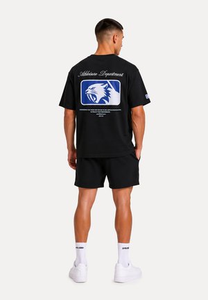 Uomo con t-shirt nera con logo di gatto selvatico blu e bianco e testo sul retro, pantaloncini neri, calzini bianchi e scarpe da ginnastica bianche, in piedi di spalle.