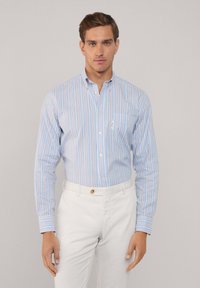 Camisa de botones azul claro con rayas verticales en varios colores, que cuenta con un bolsillo y mangas largas, combinada con pantalones blancos.