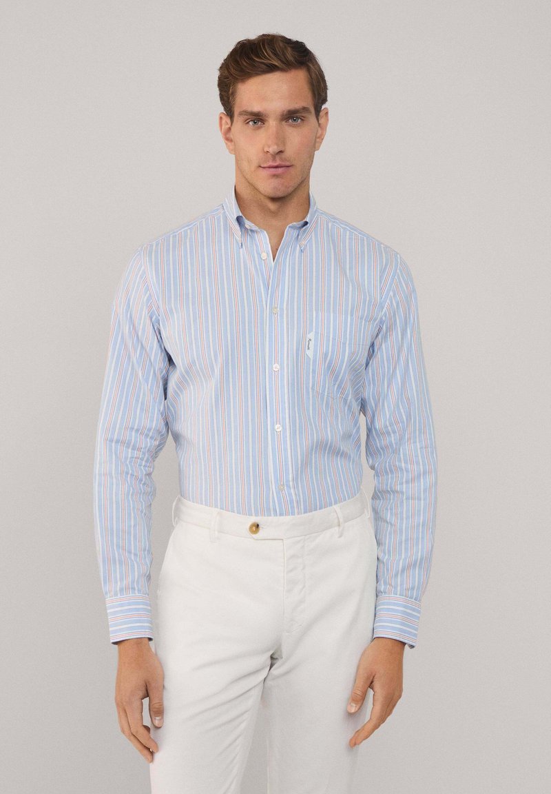 Camisa de botones azul claro con rayas verticales en varios colores, que cuenta con un bolsillo y mangas largas, combinada con pantalones blancos.