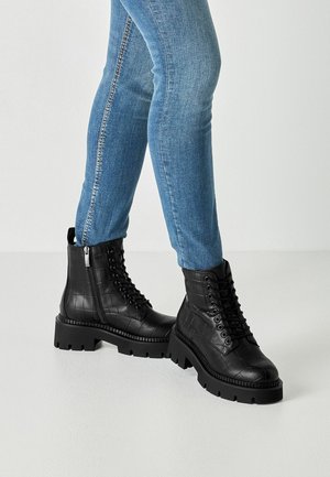 Jambes en jean bleu slim portant des bottes noires épaisses à lacets avec fermeture éclair latérale et motif texturé sur un sol blanc.