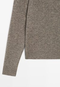 Close-up van een bruine gebreide trui mouw en onderste zoom met geribde manchetten op een witte achtergrond.