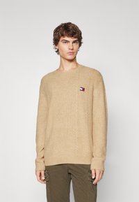 Pull en maille beige avec des poignets et un ourlet côtelés. Présente un petit patch logo multicolore sur la poitrine. Col rond classique.