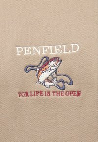 Μπεζ ύφασμα με κεντημένο το κείμενο "PENFIELD" σε λευκό χρώμα πάνω από πολύχρωμο σχέδιο ψαριού, με το κείμενο "FOR LIFE IN THE OPEN" σε κόκκινο χρώμα από κάτω.
