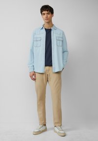 Lichtblauwe denim overhemd met twee voorzakken, gedragen over een marineblauwe t-shirt, gecombineerd met beige broeken en witte sneakers met grijze accenten.