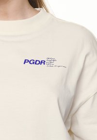 Krátký rukáv, krémové bavlněné tričko s modrým logem a textem "PGDR". Kulatý výstřih a jemná textura. Jednoduchý, čistý design bez vzorů.