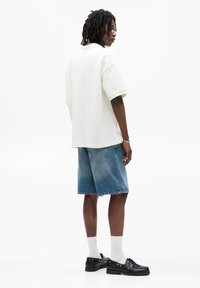 Camicia bianca oversize a maniche corte con texture, abbinata a pantaloni corti in denim blu, scarpe nere e calzini bianchi, vista di lato.