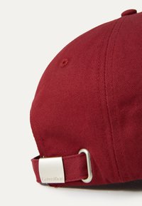 Bordeauxkleurige pet met een gebogen rand, gemaakt van textuurstof. Bevat een zilveren metalen gesp met het Calvin Klein-logo op de verstelbare band.