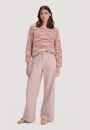 Frau trägt einen rosa-weiß gestreiften Strickpullover, weite hellrosa Hosen und beige strukturierte Slip-on-Schuhe, steht vor einem einfarbigen Hintergrund.