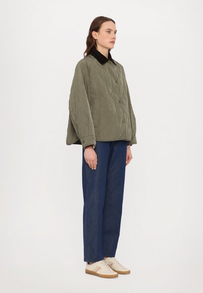 Olivgrüne gesteppte Jacke mit schwarzem Kragen, kombiniert mit blauen Denim-Hosen und beigen Schuhen mit mehrfarbigen Streifen an den Seiten.