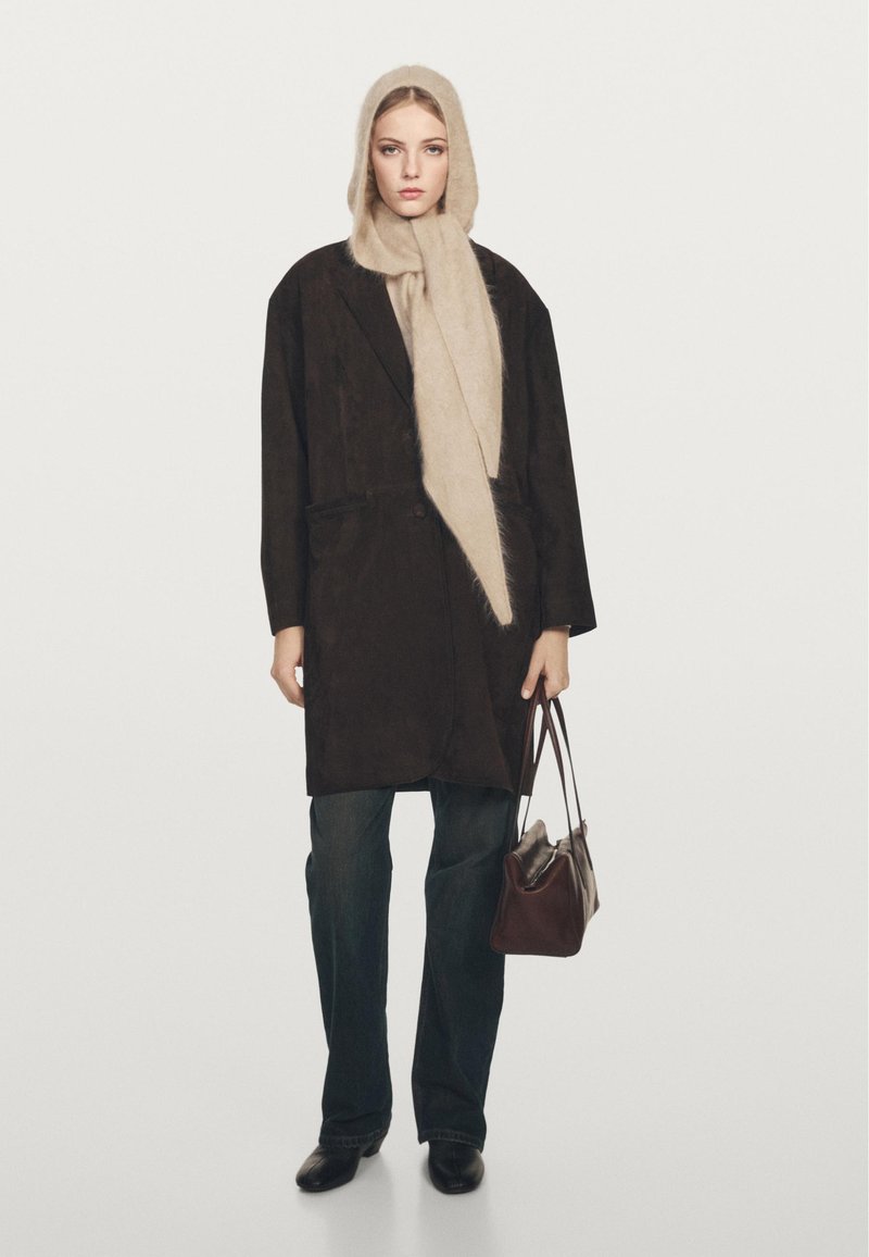 Brauner Wildledermantel mit tiefem V-Ausschnitt, beiger Schal, dunkle Jeans und schwarze Schuhe; trägt eine braune Ledertasche.