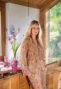 Beige loungewear met bloemmotief en levendige borduursels, bestaande uit een losvallende top en broek. Houten keukenachtergrond en natuurlijk licht.