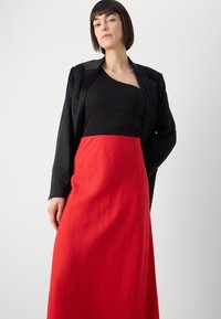 Mustmustest mustriidest bleizer musta ribatud topi ja punase põrandani ulatuva seeliku peal. Riietusel on struktureeritud disain, millel on sujuvad jooned ja minimaalne detail.