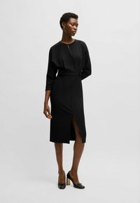 Svart midi-klänning med hög krage, långa ärmar, midjebälte och framträdande slits. Slät tyg med skräddarsydd passform. Bärs med svarta klackar.