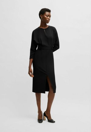 Black midi dress with a high neckline, long sleeves, waist belt, and front slit. Smooth fabric with a tailored fit. Worn with black heels.