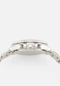 Montre en acier inoxydable avec un boîtier rond, une lunette texturée, deux boutons et un logo sur la couronne. Bracelet argenté avec maillons.