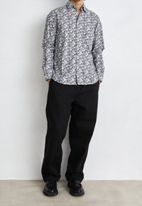 Svartvit mönstrad skjorta med knappar och spridd krage, kombinerad med lösa svarta byxor och svarta skor, allt mot en neutral bakgrund.