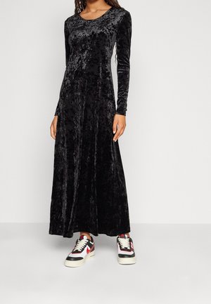 Personne portant une longue robe en velours noir froissé avec manches longues, associée à des baskets noires, blanches et rouges, debout devant un fond uni.
