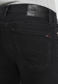 Jeans de denim negro con un acabado texturizado, que incluyen un parche de cuero con la etiqueta "TOMMY JEANS" y costuras decorativas en los bolsillos traseros.