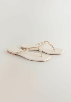 Płaskie sandały o kwadratowym kształcie noska, wykonane z miękkiego, jasnobeżowego materiału. Dwie cienkie paski krzyżują się na palcach, z węzłem jako akcentem.