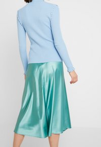 Rippiges, hellblaues Oberteil mit hohem Kragen und Rüschenbesatz; kombiniert mit einem glänzenden, türkisfarbenen Satin-Midi-Rock, der elegant fällt.