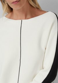 Vrouw in een witte trui met een zwarte verticale lijn in het midden en zwarte mouwen, met een close-up van de bovenste torso.