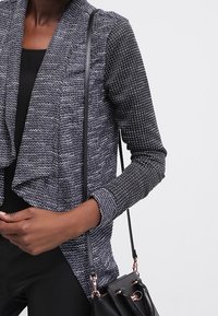 Svart och gråmönstrad cardigan med draperad front, långa ärmar och textilstruktur. En svart handväska bärs på axeln.