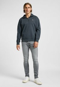 Felpa zip grigia scura con polsini a coste, accento logo discreto sul petto, abbinata a jeans grigi chiari e sneakers bianche.