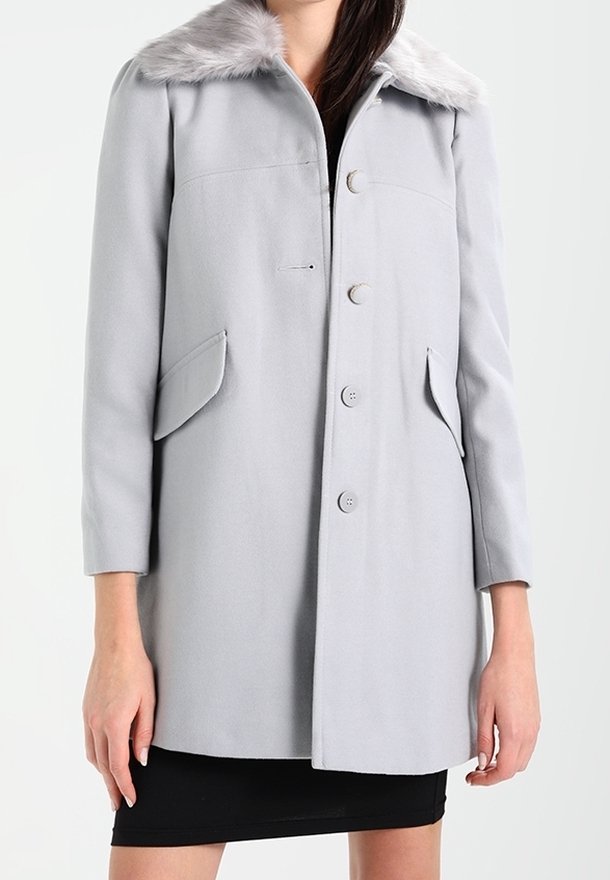 Femme portant un manteau gris clair boutonné avec des poches à rabat et un col en fourrure, par-dessus une robe noire.