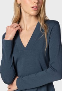 Laivastonsininen pitkähihainen toppi, jossa on V-pääntie ja tekstuuri. Mallilla on päällä geometriset hopeiset korvakorut, neutraalilla taustalla.