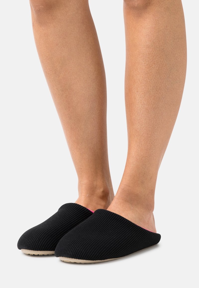Personne portant des chaussons noirs en tissu côtelé à enfiler avec des semelles beiges, visible des mollets jusqu'aux pieds sur un fond uni.