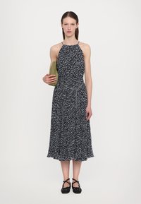Musta kukkakuosi midi-mekko ohuilla olkaimilla, istuvalla bodysuunnittelulla ja levenevällä hameella. Malli pitää kädessään vihreää laukkua ja pukeutuu mustiin kuviollisiin ballerinoihin.