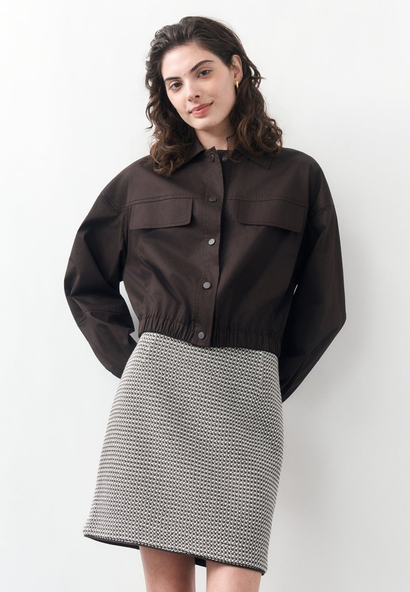 Νεαρή γυναίκα με κυματιστά μαλλιά μέχρι τους ώμους, φόραει ένα σκούρο cropped μπουφάν και μια ασπρόμαυρη φούστα με μοτίβο, μπροστά από ένα απλό φόντο.