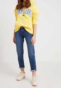 Felpa gialla con cappuccio e la scritta "TOMMY JEANS" in lettere blu, abbinata a jeans blu scuro arrotolati e sneakers beige chiaro.