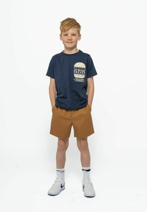 Marinblå bomullströja med grafiken "Just Say Cheese", ihop parat med bruna shorts. Vita sneakers med blå detaljer, ankelsockor med svarta ränder.