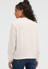 Cremefarbene, strukturierte Sweatshirt mit langen Ärmeln und rundem Ausschnitt. Getragen über blauen Jeans. Schlichtes und tailliertes Design.