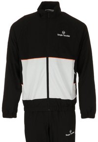 Zwart-wit trainingspak met een ritsjack, oranje accenten en logo. Heeft elastische manchet en tailleband, gladde stoftextuur.