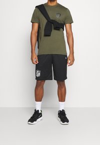 Olivgrön t-shirt med en logotyp, svarta meshshorts med ett NFL-emblem och svarta sneakers. Vita strumpor fulländar den athletiska outfiten.