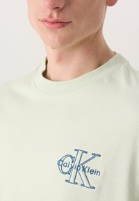 Világoszöld pamut póló, kerek nyakkivágással, a bal oldalon kék hímzett "Calvin Klein" logóval. Sima textúra.