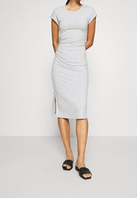 Femme portant une robe midi à manches courtes gris clair froncée avec une fente sur le côté et des sandales noires ouvertes à bout découvert, se tenant contre un fond uni.