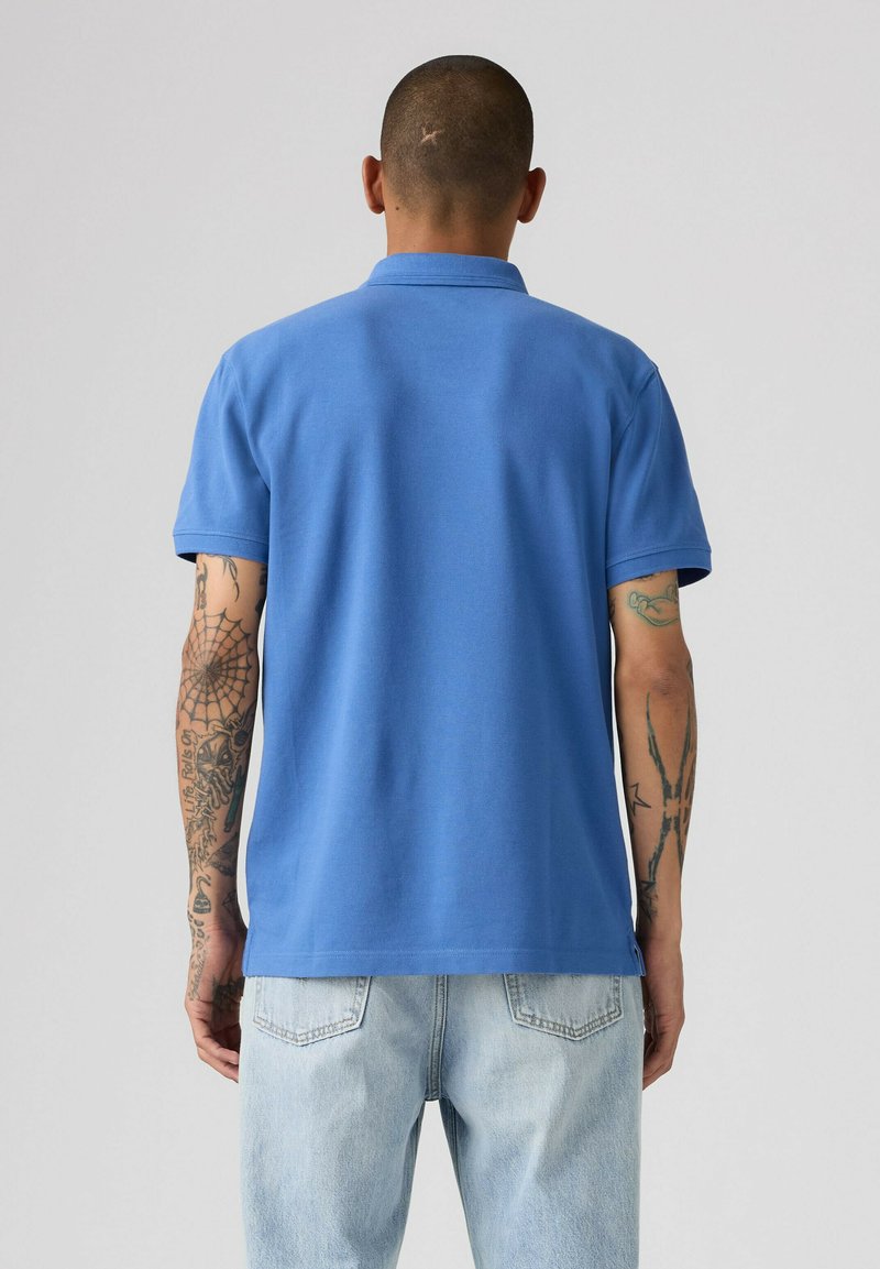 Hombre con tatuajes en ambos brazos, vistiendo una camisa polo azul de manga corta y vaqueros azul claro, de pie de espaldas sobre un fondo gris liso.
